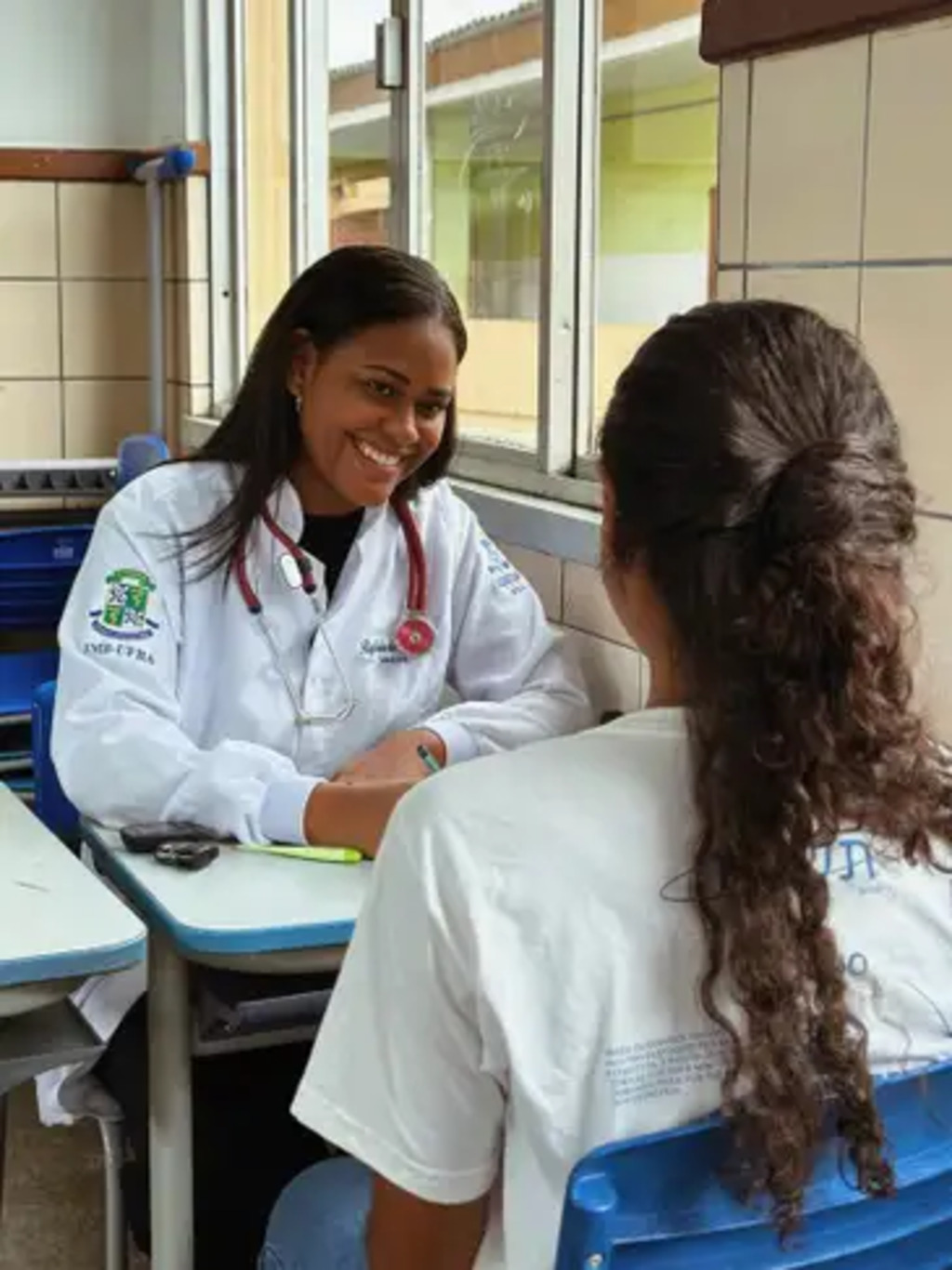 Rafaela da Silva Souza, estudante de medicina na Universidade Federal da Bahia (UFBA) e moradora de Salvador, atende uma paciente com sorriso acolhedor, vestida com jaleco branco em ambiente escolar adaptado para atendimento. Foto: Rafaela da Silva Souza/Arquivo pessoal.